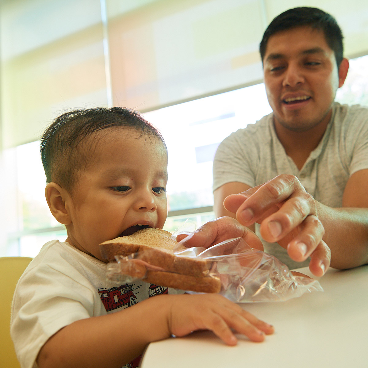 Young boy eating a sandwich with some help from this father